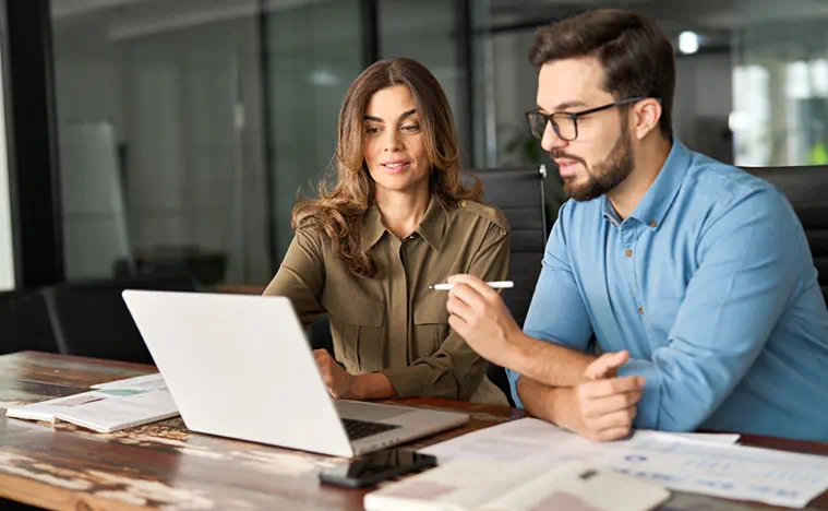 Two busy colleagues working together talking using laptop in office. Middle aged Latin female manager teaching young male worker looking at computer discussing business plan at office meeting.